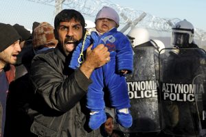 A stranded migrant holding a baby shouts next to a Greek police cordon following scuffles at the Greek-Macedonian border, near the village of Idomeni - TA NEA - TA NEA