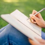Person writes in an open notebook while sitting on green grass, outdoors, with a silver pen and bracelets on the wrist.