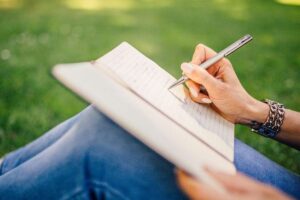 Person writes in an open notebook while sitting on green grass, outdoors, with a silver pen and bracelets on the wrist.