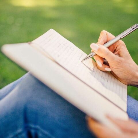 Person writes in an open notebook while sitting on green grass, outdoors, with a silver pen and bracelets on the wrist.