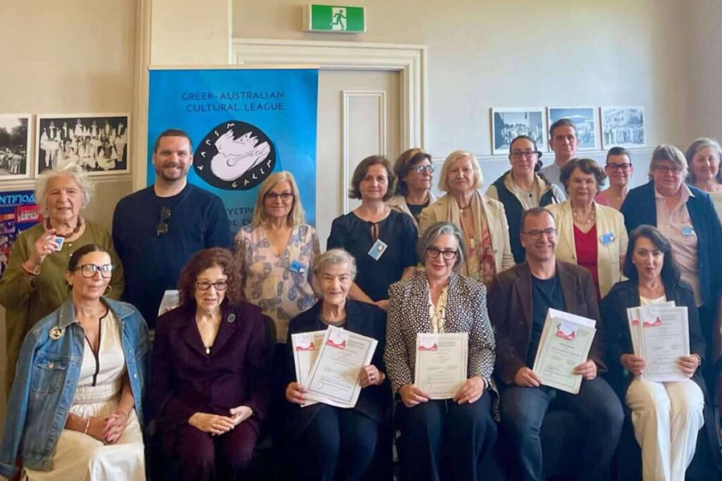 Group of people indoors posing for a photo at a Greek-Australian Cultural League event; several hold certificates.