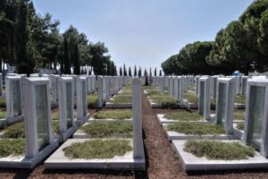 Rows of identical white tombstones in a cemetery, with a central dirt path and trees lining the grounds under a bright blue sky.