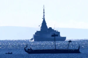 Silhouetted military ship with a tall mast on a shimmering sea, seen from the side, with a long, rowed boat in the foreground and small figures onboard.