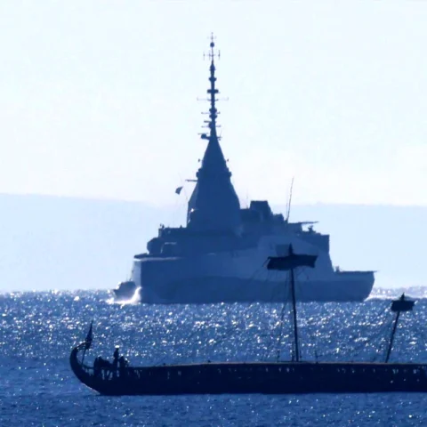 Silhouetted military ship with a tall mast on a shimmering sea, seen from the side, with a long, rowed boat in the foreground and small figures onboard.