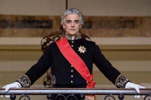Man in a black ceremonial uniform with gold embroidery, white gloves, a red sash, standing at a balcony railing.