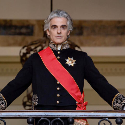 Man in a black ceremonial uniform with gold embroidery, white gloves, a red sash, standing at a balcony railing.