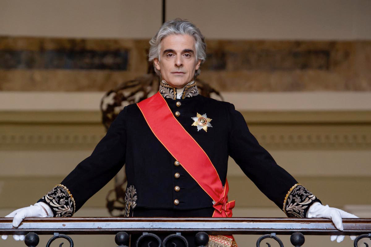 Man in a black ceremonial uniform with gold embroidery, white gloves, a red sash, standing at a balcony railing.