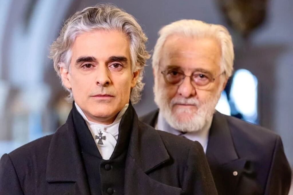 Two men in formal black coats pose for a photo, the foreground man with silver hair and a white cravat featuring a cross pin, the older bearded man with glasses behind him.