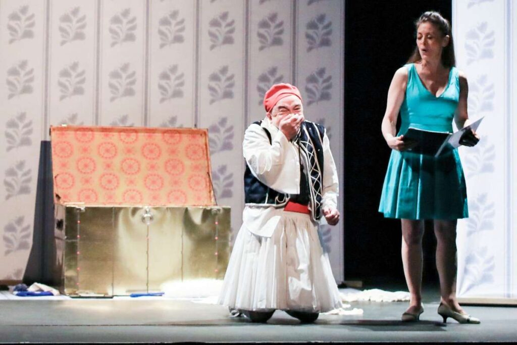 Stage scene with a man in traditional white and black costume and red fez, bowing or covering his mouth, beside a woman in a teal dress holding papers.