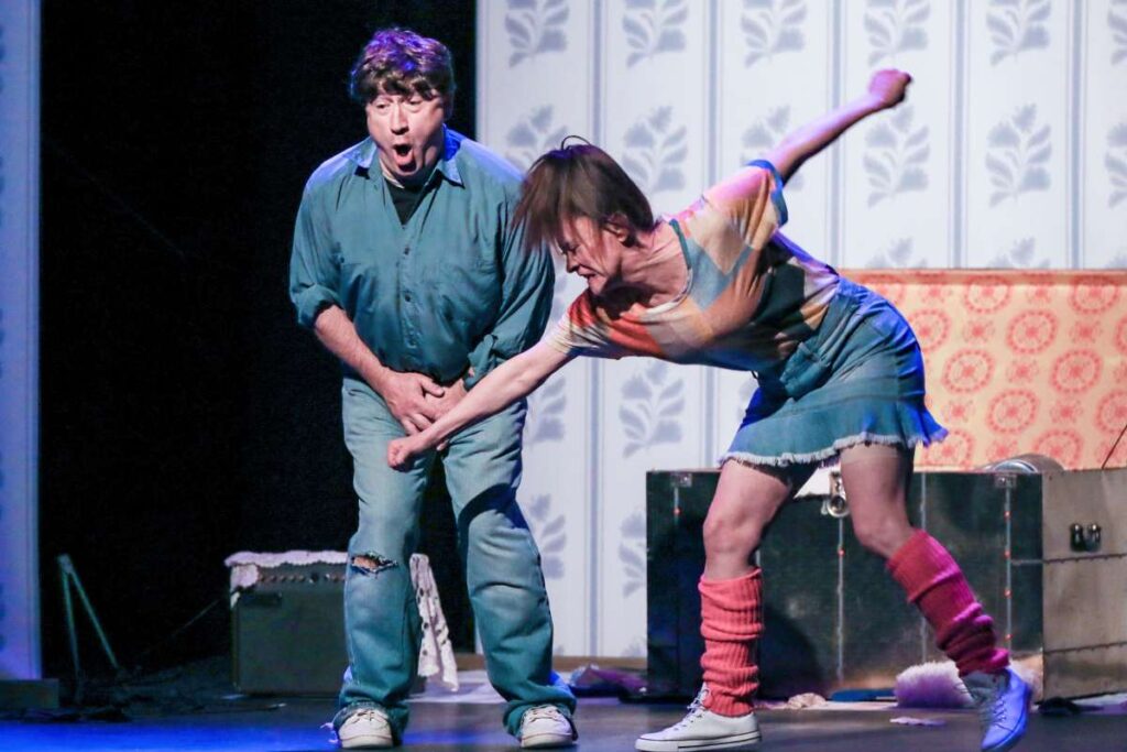 Two performers on a stage: a surprised man in a teal shirt and jeans stands as a woman in a colorful top and skirt lunges forward, reaching toward him with an extended arm amid a staged living-room backdrop.