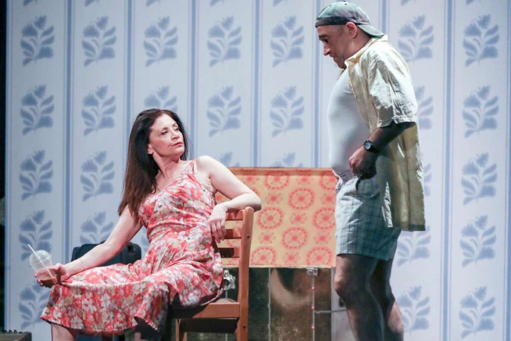 Woman in a pink floral dress sits on a wooden chair holding a cup with a straw, while a man in a backward cap and shorts stands beside her on a stage.