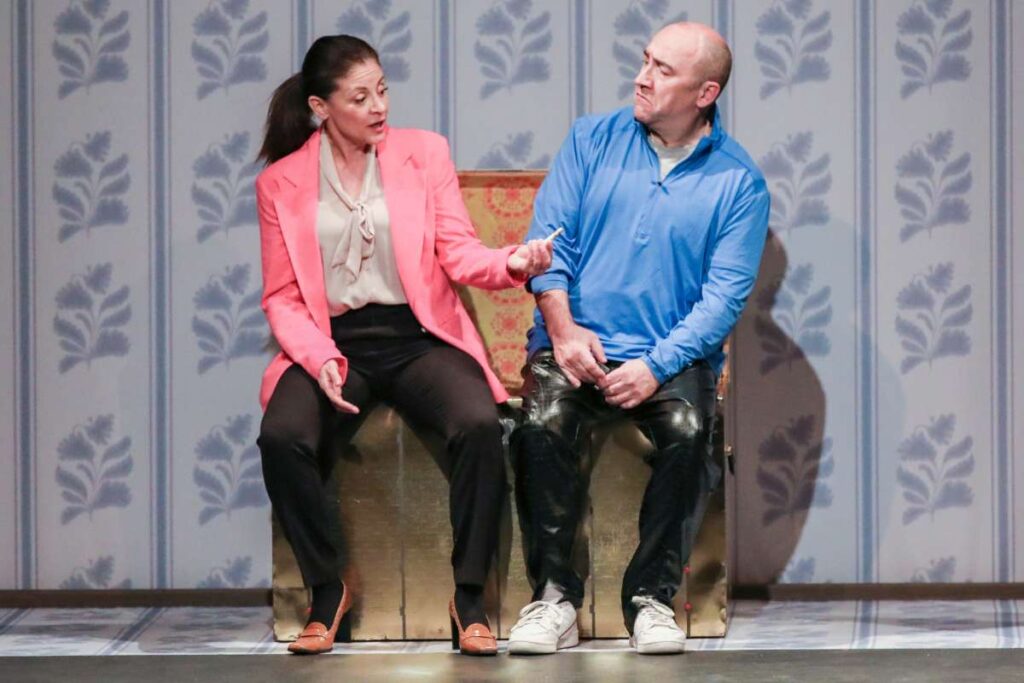 Woman in a pink blazer and beige blouse talks to a bald man in a blue shirt while seated on a gold box on stage.