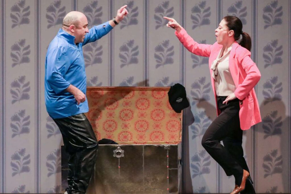Two performers on a stage point at each other, standing beside an open vintage chest with patterned lining behind them.