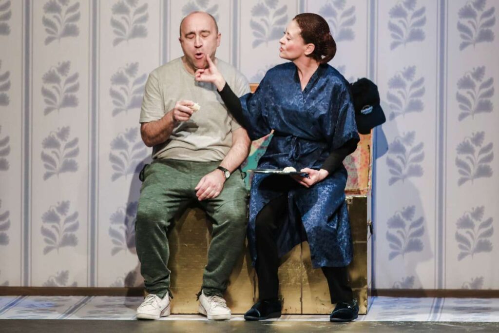 Stage scene with a woman in a blue robe gesturing with a finger toward a man’s mouth as he tastes a snack, seated on a gold prop box.