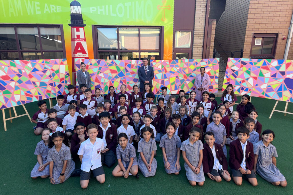 Group of students in uniform posing for a photo on a green outdoor area, with teachers in the back and a colorful mosaic wall behind them.