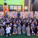 Group of students in uniform posing for a photo on a green outdoor area, with teachers in the back and a colorful mosaic wall behind them.