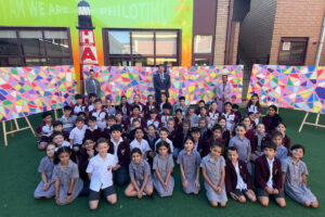Group of students in uniform posing for a photo on a green outdoor area, with teachers in the back and a colorful mosaic wall behind them.