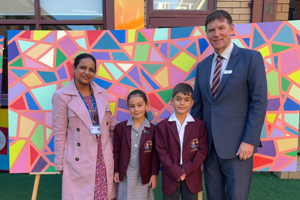 Four people posing in front of a bright geometric mural; two adults in professional attire and two students in maroon blazers with badges, outdoors at a school.