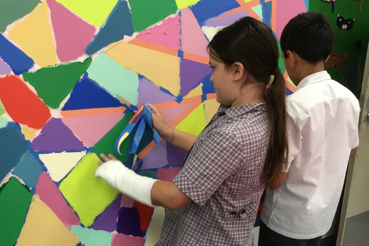Two children work on a bright, abstract mural; a girl in a plaid shirt places blue ribbons onto the colorful wall while a boy in a white shirt watches nearby.