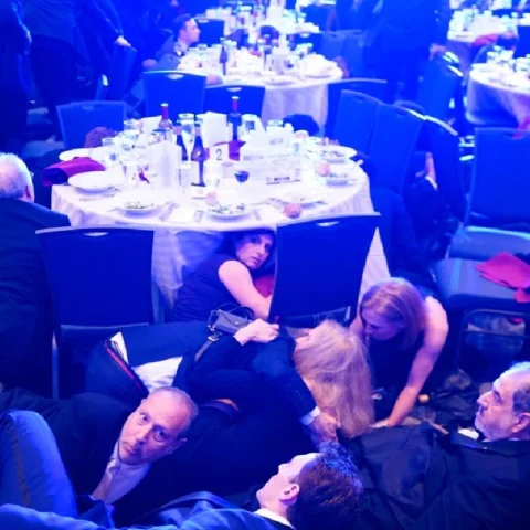 Guests crouched and hiding under a round banquet table at a gala, with plates and bottles on the table surface behind them.