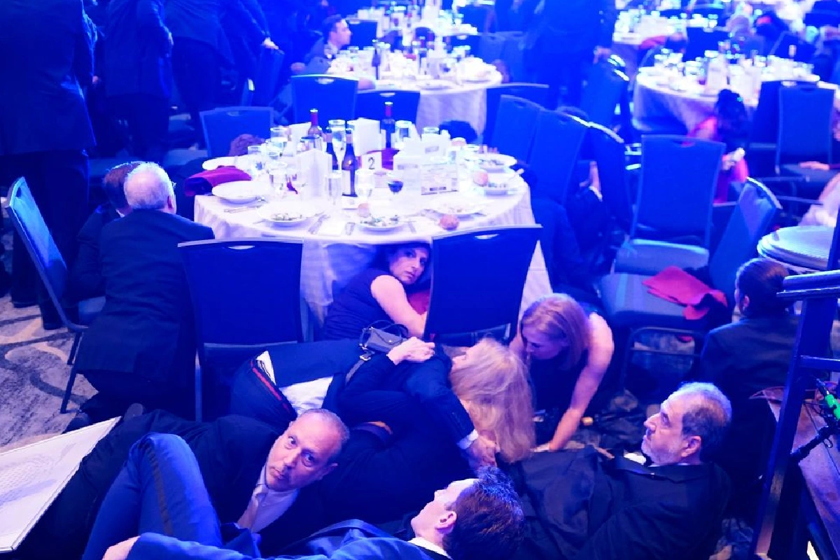 Guests crouched and hiding under a round banquet table at a gala, with plates and bottles on the table surface behind them.