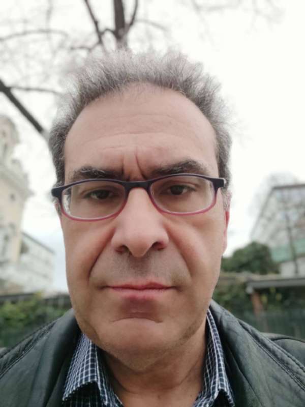 Close-up of a man with dark hair and red-framed glasses looking at the camera outdoors, neutral expression.