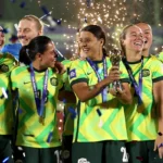 Women’s soccer team in neon yellow uniforms celebrate with a trophy and gold medals as confetti falls.