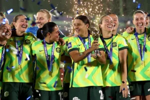 Women’s soccer team in neon yellow uniforms celebrate with a trophy and gold medals as confetti falls.