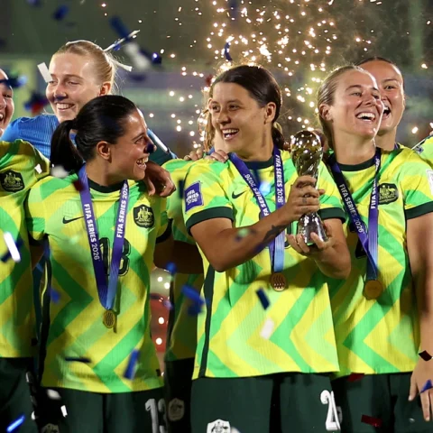 Women’s soccer team in neon yellow uniforms celebrate with a trophy and gold medals as confetti falls.