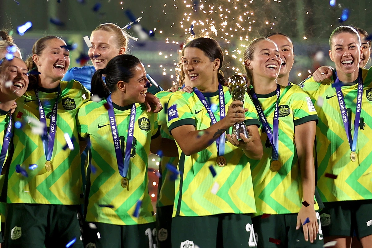 Women’s soccer team in neon yellow uniforms celebrate with a trophy and gold medals as confetti falls.