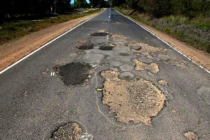 Long, straight road surface full of large potholes and missing asphalt, stretching into the distance amidst roadside trees, suggesting hazardous driving conditions.