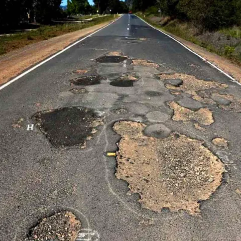Long, straight road surface full of large potholes and missing asphalt, stretching into the distance amidst roadside trees, suggesting hazardous driving conditions.