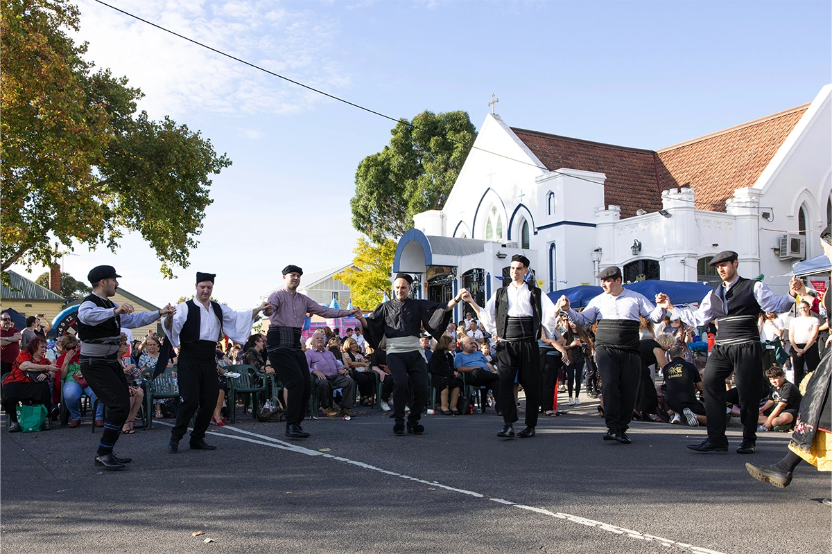 Men in white shirts and black vests dance in a circle on a sunny street while a seated audience watches from the curb, at a cultural festival in front of a white church building.