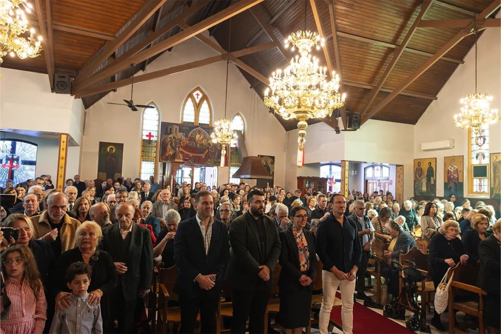 Crowded church interior with people standing for a service under ornate chandeliers under a wooden roof.