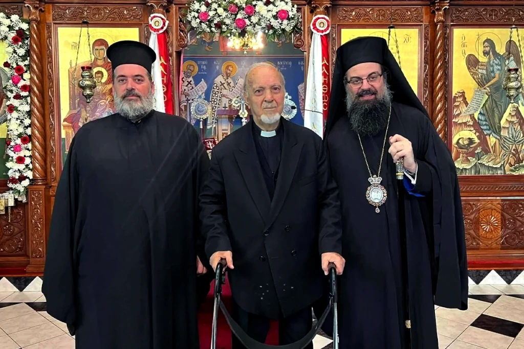 Three clergymen pose for a photo inside an ornate church with icons and flowers: a middle elderly man with a cane flanked by two priests in black robes.