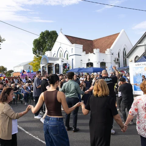 Crowd of people holding hands and dancing in a circle at an outdoor festival beside white church buildings.