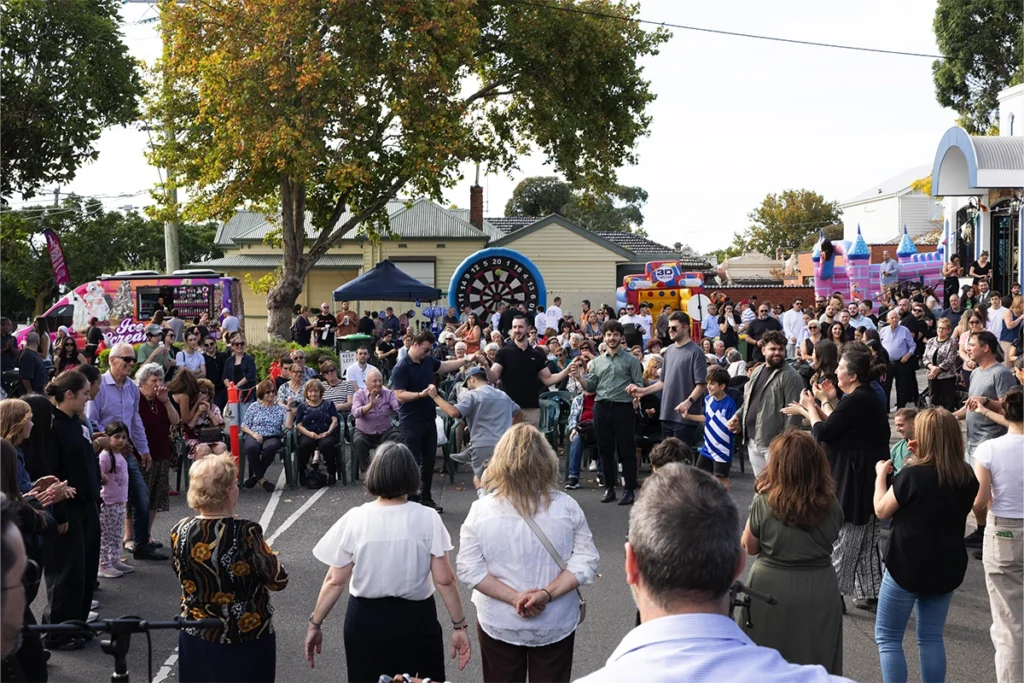 Crowd of people at an outdoor community festival with tents, a dartboard game, and inflatable rides in the background.
