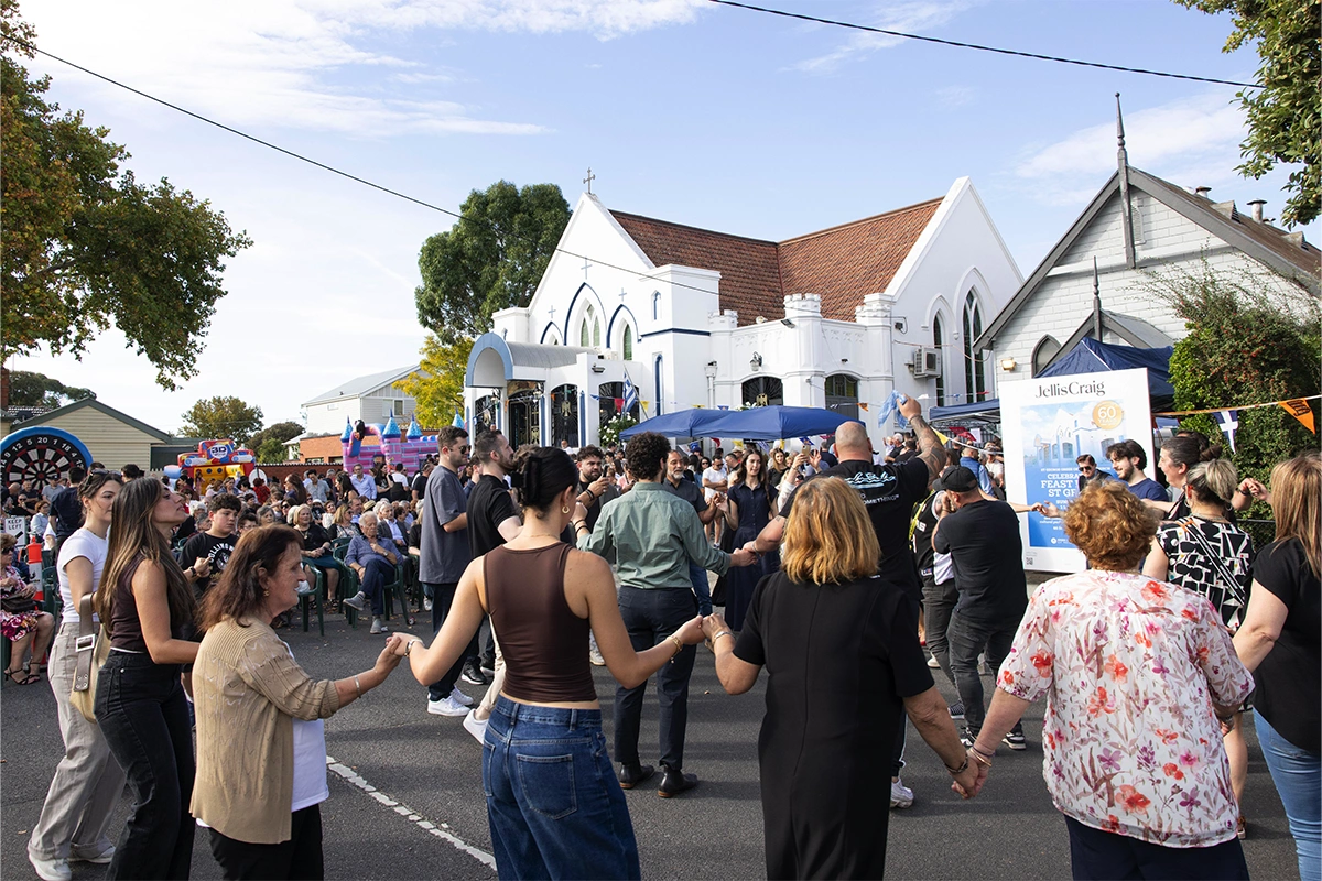 Crowd of people holding hands and dancing in a circle at an outdoor festival beside white church buildings.